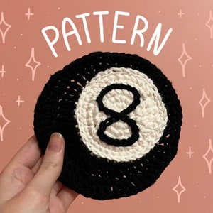May include: A handmade crochet coaster resembling an eight ball. It has a black outer ring, a white centre, and a black number eight. The word "PATTERN" is written in white script at the top.
