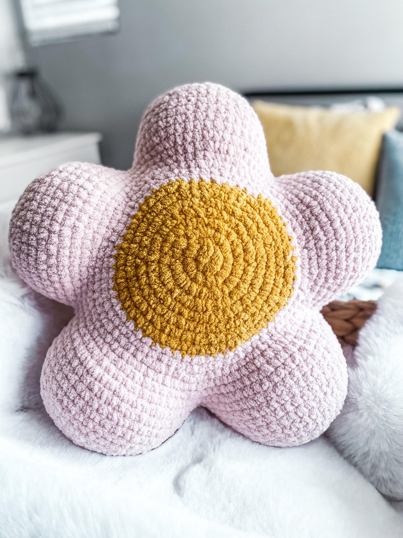 Puede incluir: Una almohada de ganchillo en forma de flor de color rosa y amarillo. La almohada tiene un gran centro amarillo y cinco p&eacute;talos rosas.