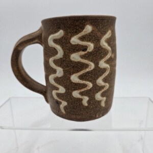 Peut inclure: Une tasse en céramique brune avec une surface texturée et mouchetée, ornée de trois lignes blanches ondulées verticales sur le devant. La tasse possède une anse incurvée sur le côté gauche et un corps cylindrique avec un bord légèrement évasé.