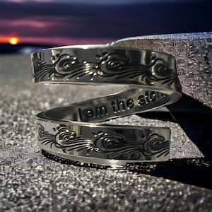 Puede incluir: Una pulsera de plata con un diseño de olas y las palabras "Soy la tormenta" grabadas en ella.