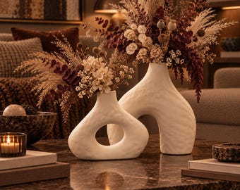 May include: Two white ceramic vases with textured surfaces and organic, asymmetrical shapes. The smaller vase features a central cutout, while the larger vase has a tall, arched form. Both contain arrangements of dried flowers in cream and deep burgundy.
