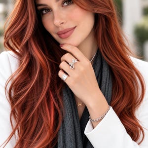 May include: A silver ring featuring a bow design with small clear stones, a wide silver band ring, a delicate gold-tone necklace with an H-shaped pendant, and a silver chain link bracelet. The jewelry is worn with a white blazer and a dark gray scarf.