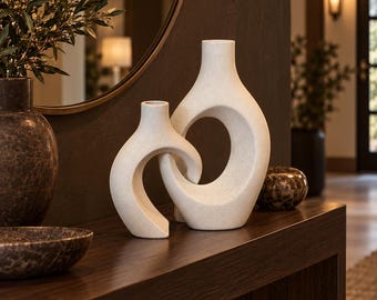 May include: Two off-white ceramic vases with a speckled texture, featuring an interlocking abstract design. The taller vase has a central circular cutout, while the smaller vase curves around it, creating a connected sculptural form.