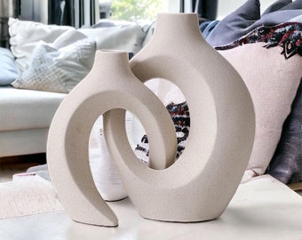 May include: Two abstract ceramic vases in a light beige color. The vases are shaped like a continuous loop with a hole in the center. The vases are sitting on a light brown table.