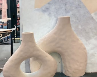 May include: Two ceramic vases in a light beige color. The vase on the left has a large oval opening in the center. The vase on the right has a rounded arch opening.