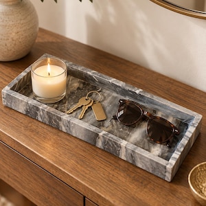 May include: A rectangular gray and white marble tray holding a glass candle with a lit white wax pillar, a gold-toned metal keyring with two keys and a blank tag, and a pair of dark brown tortoiseshell-patterned sunglasses with dark lenses.