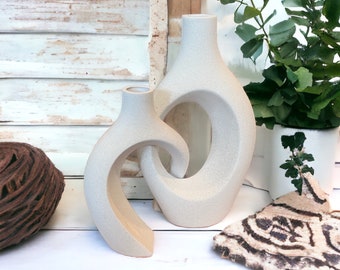 May include: Two abstract ceramic vases in a light beige color. The larger vase has a circular opening in the side. The smaller vase has a curved opening. The vases are sitting on a white surface.