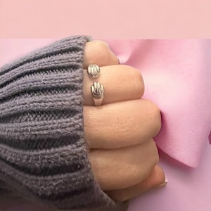 以下が含まれることがあります： 2つの手が組み合わされた銀のリング。リングは灰色のセーターを着た手の指につけられています。