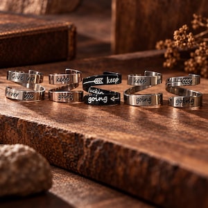 May include: Five spiral wrap rings made of silver-toned and black metal. The rings feature engraved text including phrases like Give up, never, keep going, and keep fuckin going, along with arrow symbols. Each ring has an open, adjustable design.