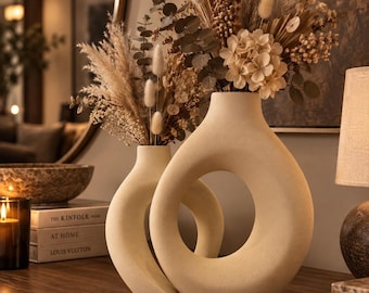 May include: Two beige ceramic vases with a smooth, matte finish and a hollow, circular center design. The vases are filled with a dried floral arrangement featuring pampas grass, eucalyptus leaves, and small white flowers in neutral tones.