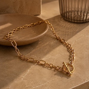 May include: A gold-toned chain necklace featuring a series of elongated oval links and a prominent toggle clasp closure. The necklace is constructed from polished metal links that form a continuous loop, secured at the front by a T-bar and ring.