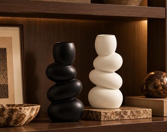 May include: Two sculptural vases, one matte black and one matte white, each featuring a stacked, pebble-like organic shape with a narrow opening at the top. These decorative vessels are displayed on a dark wooden shelf alongside books and bowls.