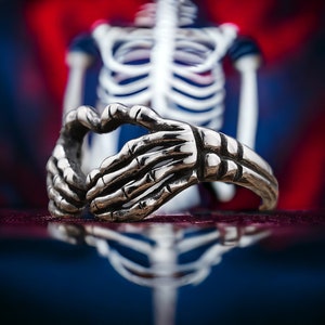 May include: A silver ring with a skeletal hand design, the hands are clasped together in a heart shape.