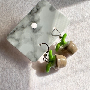 May include: A pair of dangle earrings featuring miniature potted plant charms with brown pots and bright green leaves. The earrings are attached to a white and gray marble-patterned rectangular display card with a single hole at the top center.