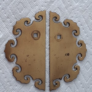 May include: Two antique brass hinges with a decorative, ornate design. The hinges are in the shape of a stylised cloud with a square hole in the centre. The hinges are worn and have a patina finish.