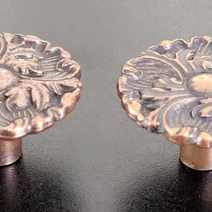 May include: Two ornate copper-coloured cabinet knobs with a floral design. The knobs have a round base and a raised, detailed floral pattern.