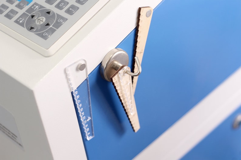 Pu&ograve; includere: Primo piano di una superficie metallica blu con tre righelli attaccati ad essa mediante magneti. I righelli sono in legno, plastica e metallo. I righelli sono di dimensioni diverse e hanno diverse marcature.