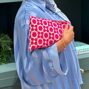 May include: A pink clutch with a white geometric pattern is held against a light blue striped shirt. The clutch has a rectangular shape and a zipper closure. The shirt has long sleeves and a button-down collar. A gold bracelet is visible on the wrist.