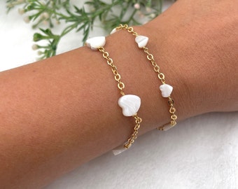 May include: A gold chain bracelet with white heart-shaped charms. The bracelet is layered on a person's wrist.