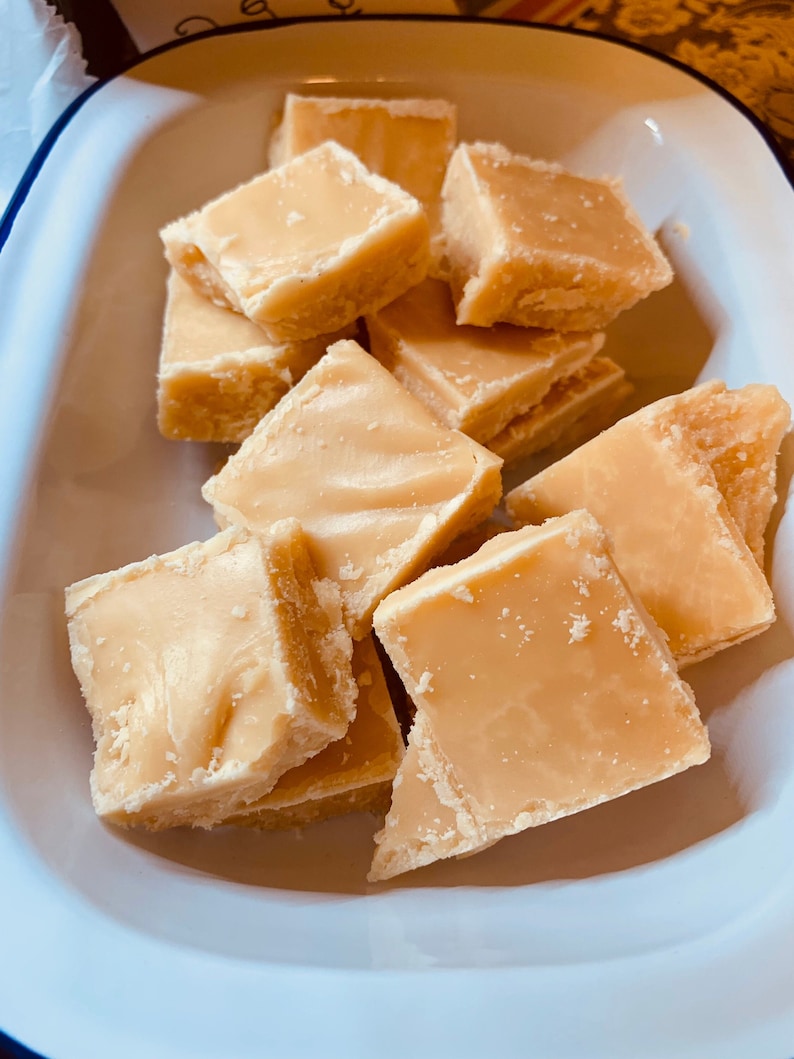 Może przedstawiać: Zbliżenie na kilka kawałk&oacute;w jasnobrązowego fudge w białej emaliowanej misce z niebieską obw&oacute;dką. Fudge jest pocięty na kwadratowe kawałki i ma gładką, błyszczącą powierzchnię. Kawałki są ułożone w stos i ułożone w misce.