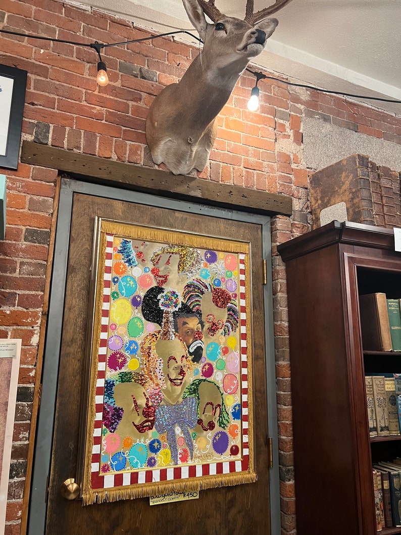 May include: A colorful beaded tapestry depicting multiple clowns with a red, white, and gold border. The tapestry is hanging on a wooden door with a price tag that reads "Beadwork Tapestry $450". A mounted deer head is above the door.