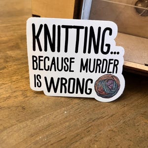 Puede incluir: Pegatina blanca con texto negro que dice "Knitting... Because murder is wrong." Una bola de hilo de colores está a la derecha del texto.