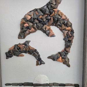 Könnte beinhalten: Gerahmtes Kunstwerk mit zwei Delfinen, die aus verschiedenfarbigen Haifischzähnen gefertigt wurden. Die Delfine sind vor einem weißen Hintergrund platziert, mit einer Sonne und einer Küstenlinie aus fossilen Materialien. Der Rahmen ist neutralgrau.