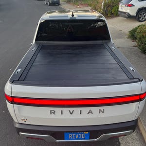 以下が含まれることがあります： 黒いトノカバーが付いた白いリビアンピックアップトラック。トラックには「RIV3D」と書かれたカリフォルニア州のナンバープレートが付いています。