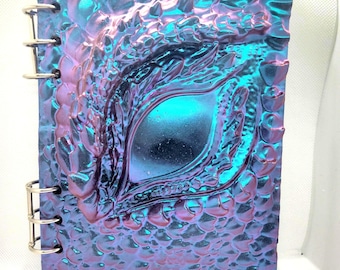 May include: A spiral bound notebook with a blue and purple iridescent cover featuring a dragon eye design.