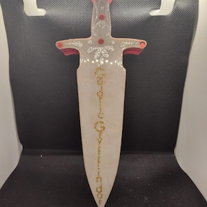 May include: A white sword with gold glitter accents and red details. The sword has the words "Gryffindor" written down the blade in gold glitter.