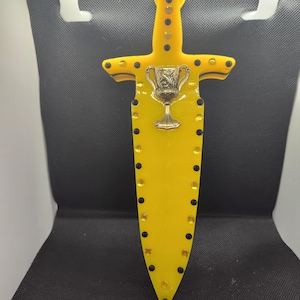 May include: A yellow plastic sword with a black handle and a gold-colored goblet on the blade. The sword is decorated with black dots and gold-colored rhinestones.