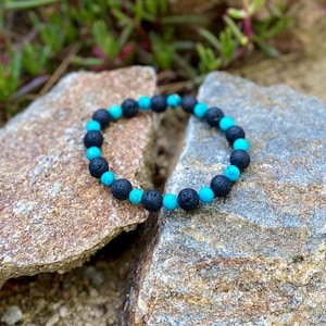 Op de afbeelding: Een zwarte en turquoise kralen armband met lavastenen kralen. De armband ligt op een stenen achtergrond.
