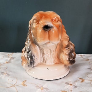 May include: A ceramic figurine of a brown and white cocker spaniel dog with a white base. The dog's head is tilted slightly to the side, and its eyes are closed.