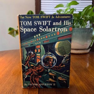May include: A vintage book titled "TOM SWIFT and His Space Solartron" with a retro space-themed cover illustration. The cover art features astronauts in space suits, a control panel, and a large moon. The author's name, Victor Appleton II, is also visible.