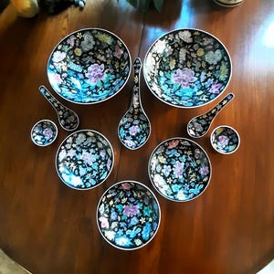 May include: A set of black and white porcelain bowls and spoons with floral designs. The bowls are arranged in a heart shape with the spoons on either side.