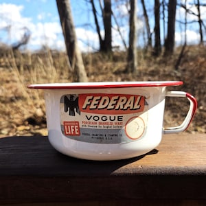 May include: White enamelware cup with a red rim and a black eagle logo. The cup has a label that reads "Federal Vogue Porcelain Enameled Ware with Titanium for Tougher Surfaces. Federal Enameling & Stamping Co. Pittsburgh, U.S.A."