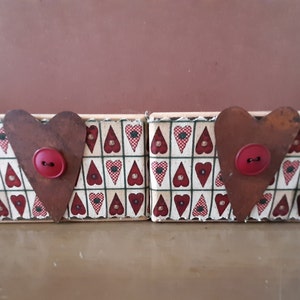 Puede incluir: Dos pequeñas cajas de madera con un diseño en forma de corazón. Las cajas están cubiertas de una tela blanca con un patrón de corazones rojos y verdes. Cada caja tiene un corazón de metal oxidado con un botón rojo en el centro.