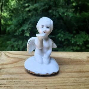 Puede incluir: Una figurita de porcelana blanca de un ángel con alas, arrodillado y soplando un beso. El ángel tiene rasgos faciales pintados y lleva un borde dorado.