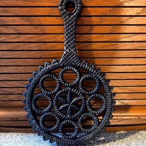 May include: Black cast iron trivet with a handle and a decorative circular design. The trivet features a star in the centre, surrounded by circles and a scalloped edge. The handle has a textured pattern.
