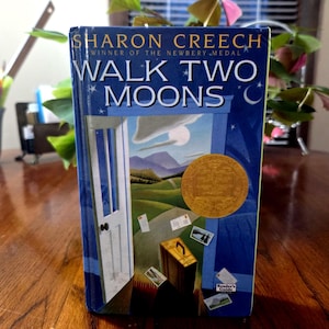 May include: A hardback book titled "WALK TWO MOONS" by Sharon Creech. The cover is blue with an illustration of a door, a landscape, and a gold medal. The book is a Newbery Medal winner.