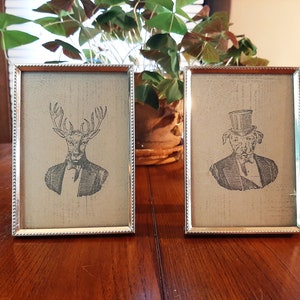 May include: Two silver picture frames with a white background. The left frame features a black and white illustration of a deer wearing a suit and bowtie. The right frame features a black and white illustration of a dog wearing a top hat and suit.