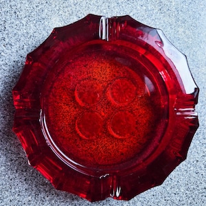 Puede incluir: Un cenicero vintage de cristal rojo rubí. El cenicero tiene una forma octogonal facetada con cuatro hendiduras circulares en el centro. El cristal tiene un aspecto moteado y es muy reflectante.