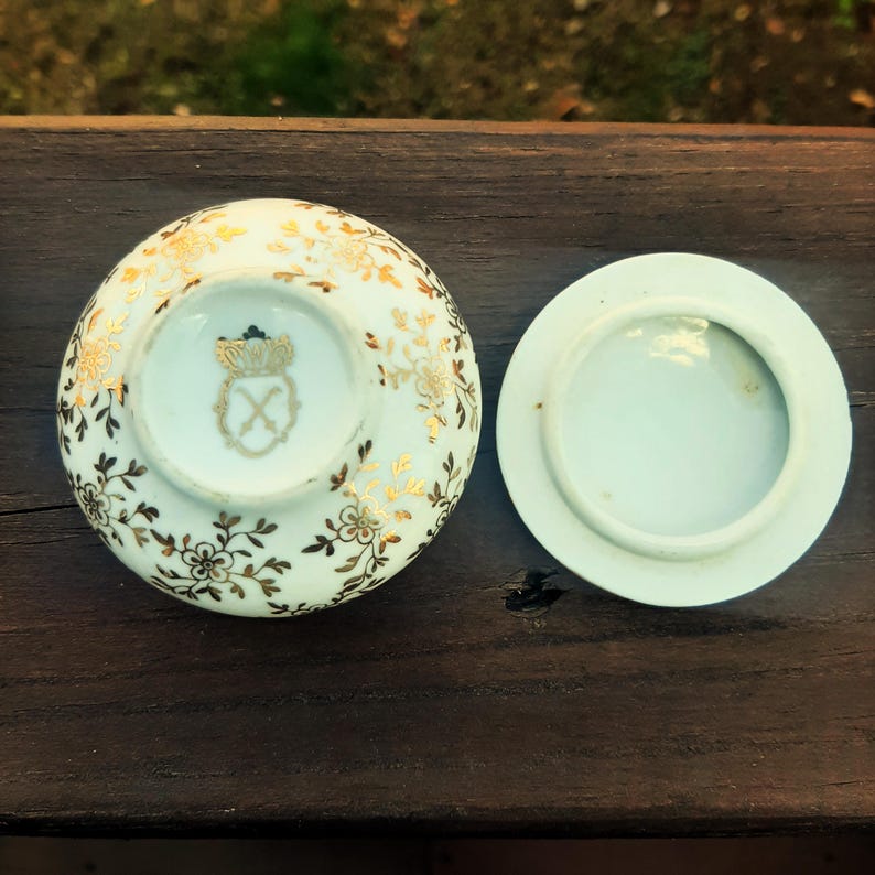 May include: A small, round, white porcelain trinket box with a separate lid. The box and lid are decorated with gold floral patterns and a gold emblem. The box is open, revealing the interior and the emblem on the base.