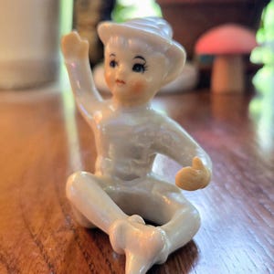 Pode incluir: Pequena figura de porcelana branca de uma criança sentada usando um chapéu. A figura tem bochechas rosadas, olhos azuis e está em pose com um braço levantado. A figura está em uma superfície de madeira.