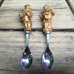 May include: Two gold angel-shaped spoon handles with silver spoons. The angels are playing musical instruments.