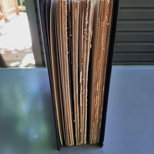 May include: A black hardback book with aged, yellowed pages. The book is closed and standing upright on a grey surface.