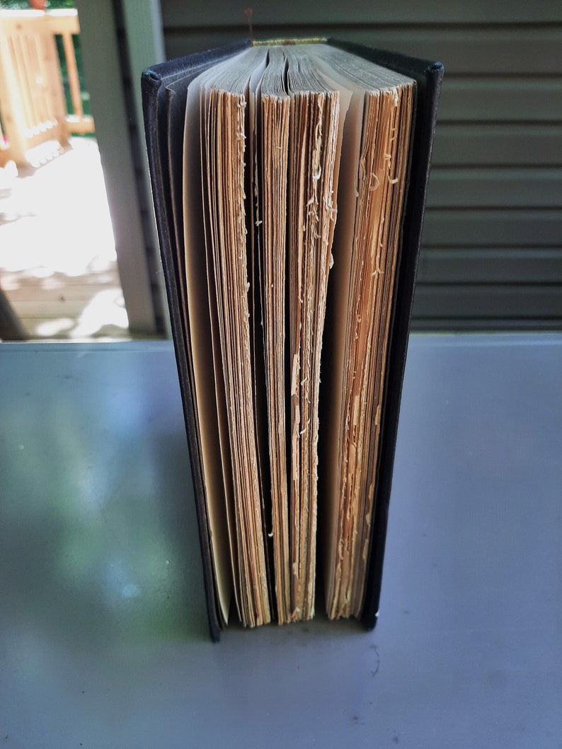 May include: A black hardback book with aged, yellowed pages. The book is closed and standing upright on a grey surface.