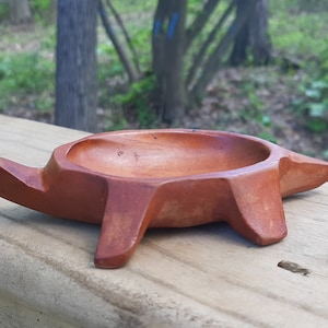 May include: A small, wooden turtle-shaped dish with a brown glaze. The dish is hollowed out and has a rounded shape. It is sitting on a wooden surface.