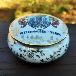 May include: A small, round, light blue ceramic trinket box with a gold rim and decorative gold floral and leaf patterns. The lid features a crest with a castle and the text "WITZENHAUSEN / WERRA".