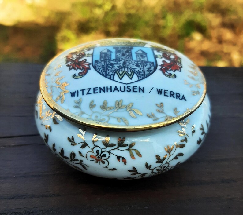 May include: A small, round, light blue ceramic trinket box with a gold rim and decorative gold floral and leaf patterns. The lid features a crest with a castle and the text "WITZENHAUSEN / WERRA".